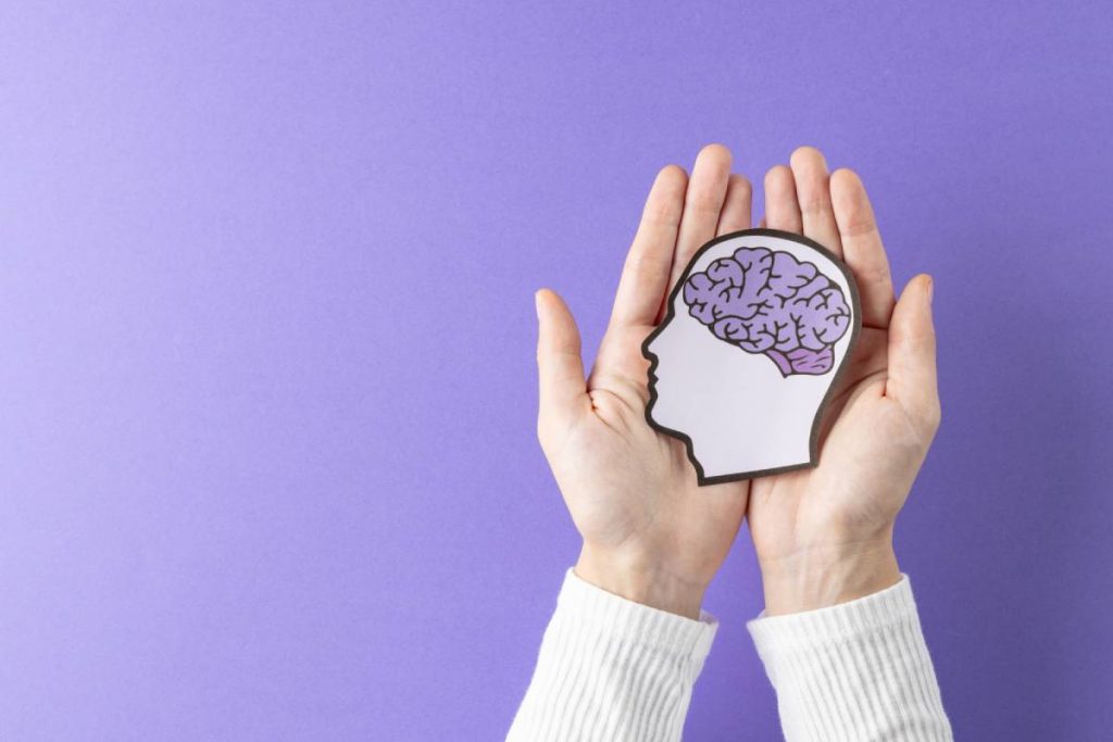 Composition of hands holding head silhouette with purple brain on purple background, with copy space. Medical services, healthcare and mental health awareness concept.