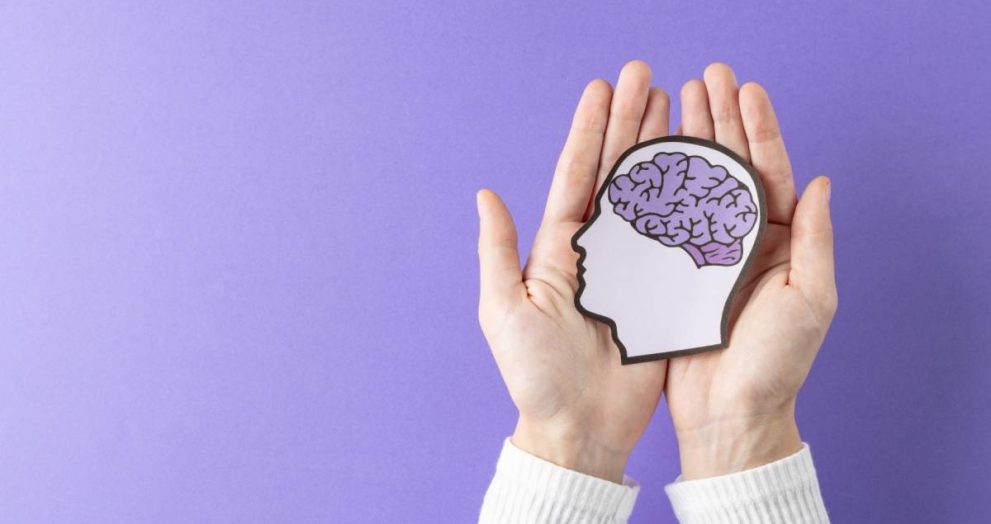 Composition of hands holding head silhouette with purple brain on purple background, with copy space. Medical services, healthcare and mental health awareness concept.