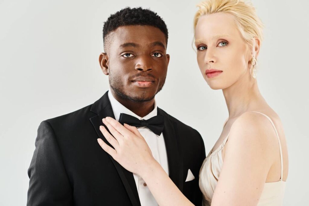 A beautiful blonde bride in a white dress stands next to an African American groom in a tuxedo, exuding elegance and love.