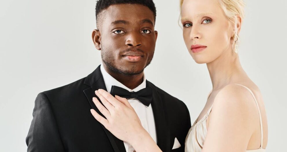 A beautiful blonde bride in a white dress stands next to an African American groom in a tuxedo, exuding elegance and love.