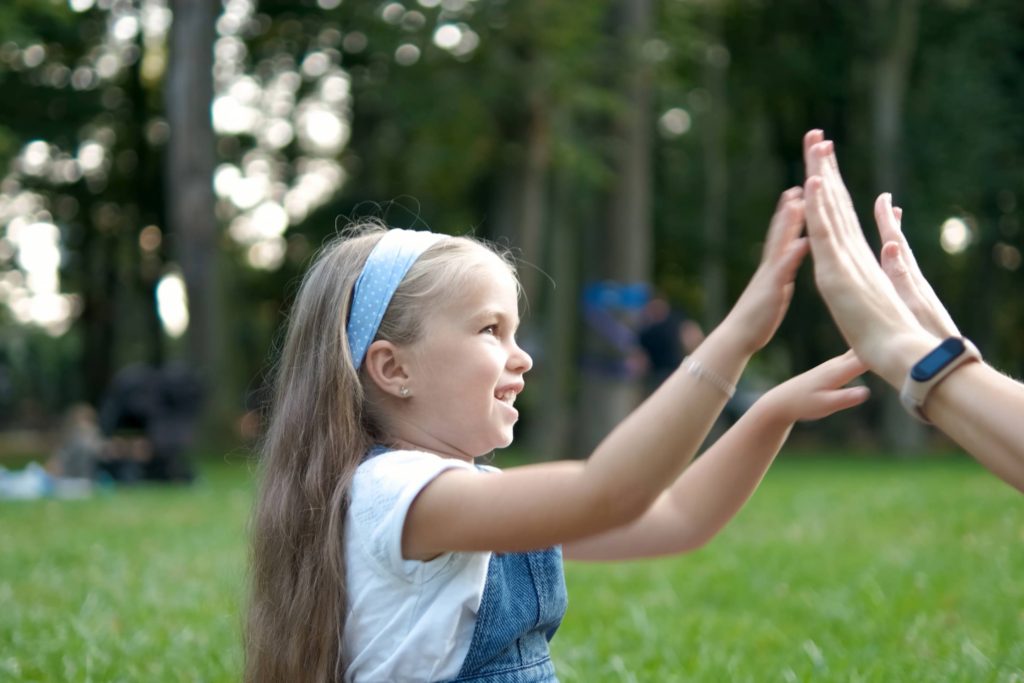 Pretty little child girl playing game with her mom clapping their hands together outdoors in green summer park.