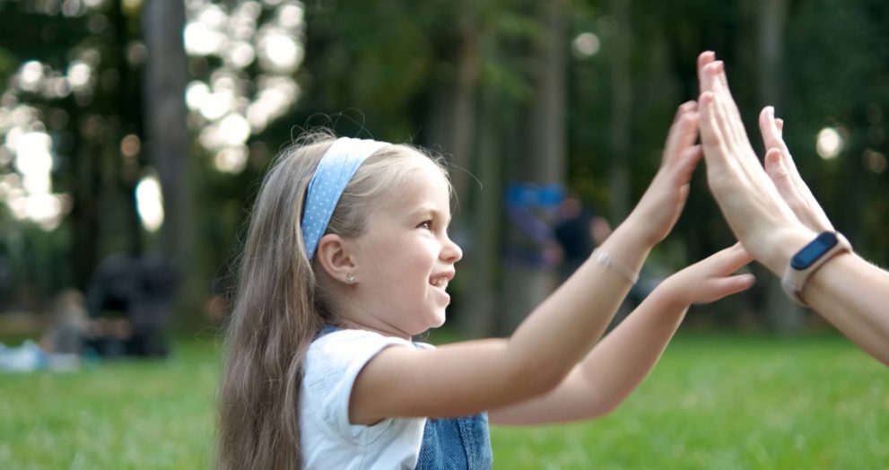 Pretty little child girl playing game with her mom clapping their hands together outdoors in green summer park.
