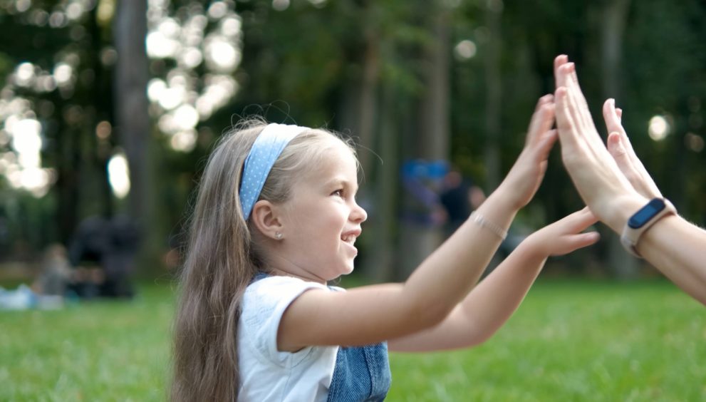 Pretty little child girl playing game with her mom clapping their hands together outdoors in green summer park.