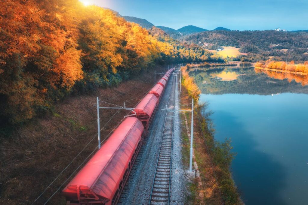 Aerial view of red freight train moving near river in alpine mountains at sunrise in autumn. Top view of wagons, railroad, lake, reflection in water, orange trees in fall. Railway station in Slovenia